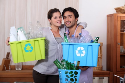A young couple standing indoors in a room with light-colored wooden paneling and a wooden cabinet in the background. They are holding large plastic recycling bins; the woman on the left has a bright green bin filled with empty glass bottles and transparent plastic containers, while the man on the right has a light blue bin containing mostly empty glass bottles with green and clear glass. In front of him is a smaller, dark blue plastic bin filled with additional empty green glass bottles. Both individuals are smiling, dressed casually, and appear to be engaged in a recycling activity typical of rubbish management services. The scene is well-lit, emphasizing the clear textures and varied colors of the containers and recycled materials, illustrating household waste sorting for disposal or recycling.