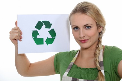 A young woman with blonde hair styled in a side braid, wearing a green shirt and a striped apron, holds up a white sign with a green recycling symbol consisting of three arrows chasing each other in a triangular loop. She is standing against a plain white background, and her expression is neutral and professional. The woman appears to be indoors, and her gesture suggests awareness of recycling practices, which aligns with the context of waste management and rubbish removal services in the Putney area, as indicated by the webpage for Putney House Clearance. The sign and the woman's attire convey a message of environmental responsibility and proper waste disposal. This image could be used to visually support content related to environmentally conscious rubbish removal and house clearance services offered by Putney House Clearance in Putney, SW15, or nearby areas.