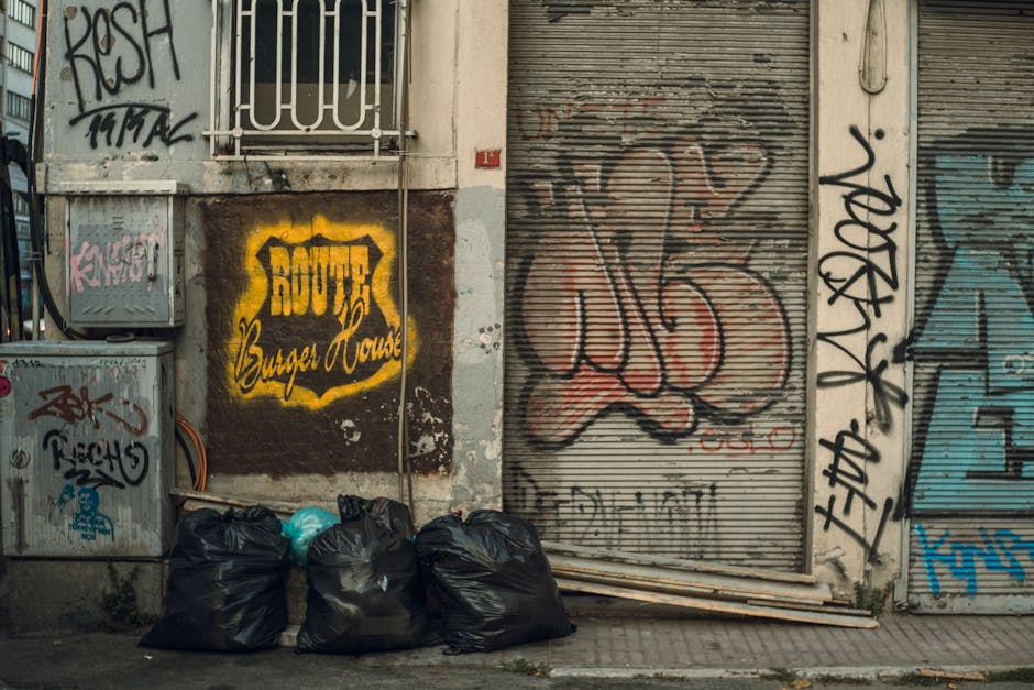 Five black rubbish bags filled with waste are lined up on a sidewalk in front of a wall covered with various graffiti tags and safety barrier pipes. The bags appear to be full and tightly sealed, rest