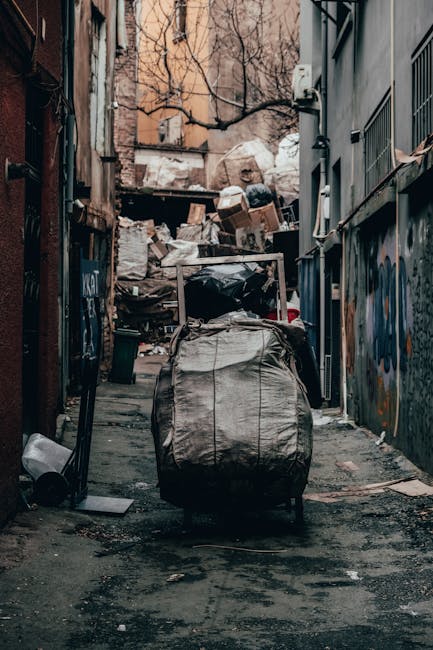 Five black rubbish bags filled with waste are lined up on a sidewalk in front of a wall covered with various graffiti tags and safety barrier pipes. The bags appear to be full and tightly sealed, rest