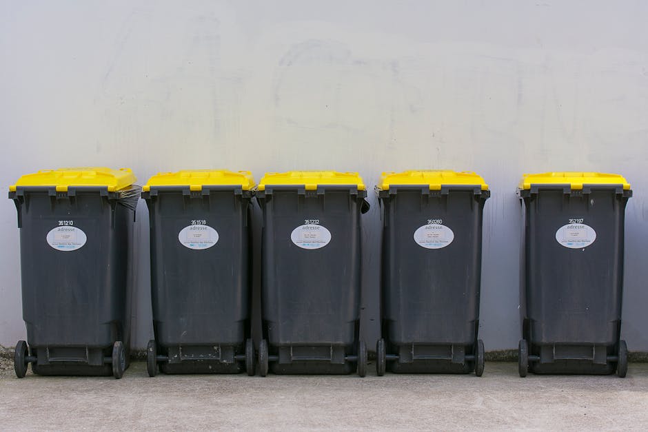 A pile of discarded cardboard boxes, some flattened and others still in their original shape, along with plastic packaging and a woven sack, are stacked at the base of a brick wall outside a building.